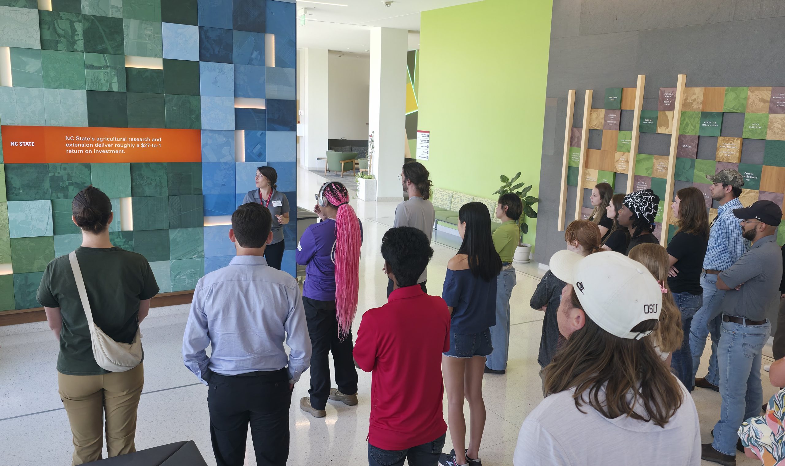 A group of people stand in a building lobby for a tour. A woman in the front is the tour guide, standing in front of an interactive wall of blue and green panels.